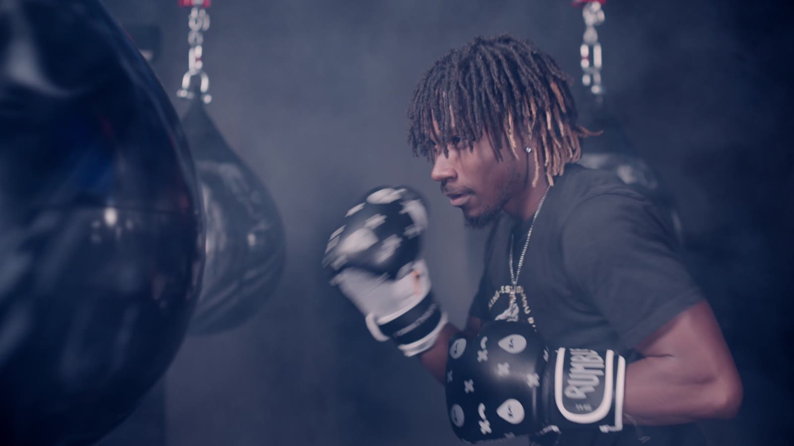 A boxer with locs throwing a punch beside a heavy bag in a smoky gym