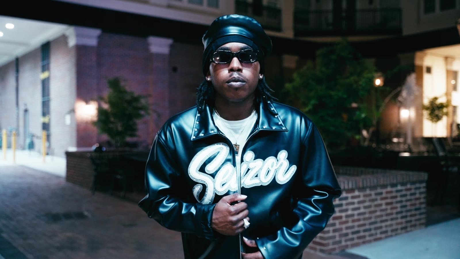 A model in a black leather varsity jacket and sunglasses in a brick courtyard at night