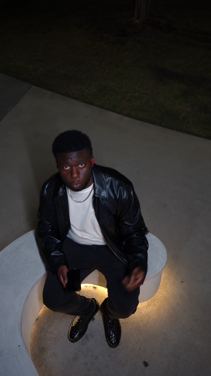 Artist Elisha in a leather jacket seated under a light at night
