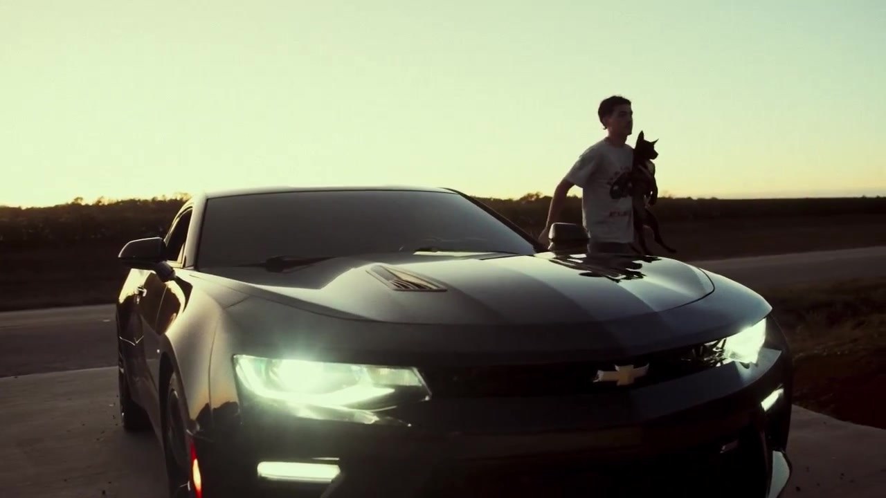 A black Camaro with headlights on at sunset, its owner holding his dog beside it