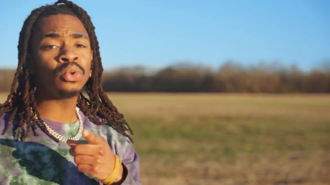 Omri in a tie-dye sweatshirt rapping to camera in an open field under a blue sky